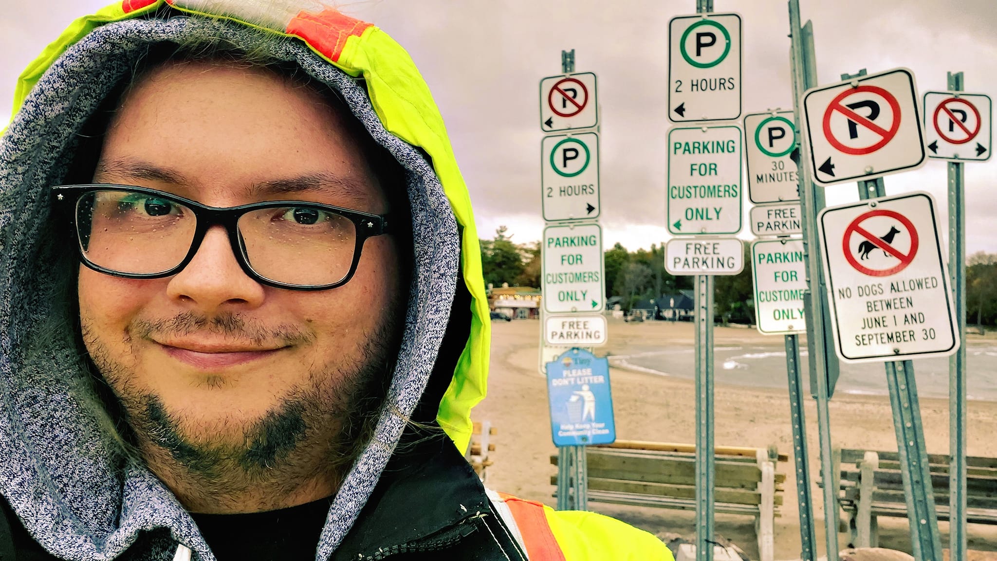 A man with long green hair wearing is wearing a high visibility raincoat in front of several parking signs, which have been stored together for the winter